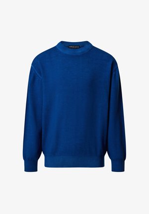 Garçon debout portant un pull bleu, une chemise rayée, un short cargo beige, des chaussettes blanches et des baskets blanches sur un fond uni.