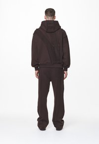 Chándal marrón con sudadera de gran tamaño y pantalones de corte relajado. Presenta costuras texturizadas y un cuello alto. Diseño minimalista, sin logotipos visibles.