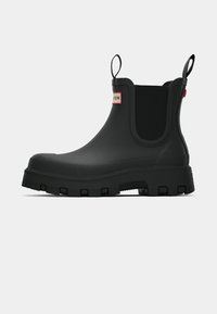 Botas Chelsea de goma negra con una superficie texturizada, paneles laterales elásticos y una suela gruesa y robusta. Etiqueta de marca roja en la parte superior.