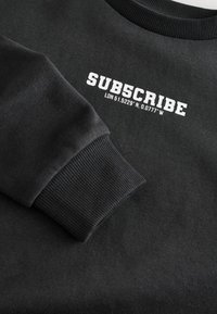 Sort sweatshirt med ribbede manchetter og halsudskæring. Forsiden har hvid tekst: "SUB5CRIBE LDN 51.5229° N. 0.0777° W", vist fremtrædende.