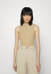 Top de gola alta sem mangas em bege com textura canelada, combinado com calças largas em tom creme, com bolsos frontais e fecho de botão.