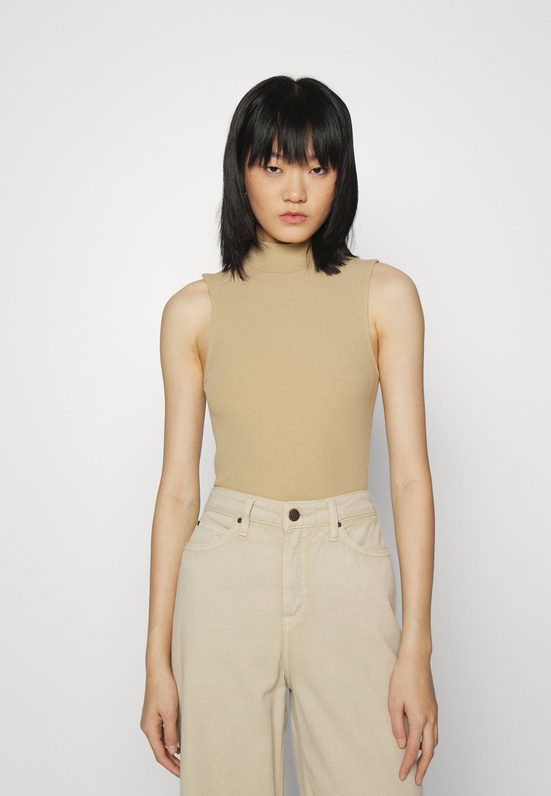 Top de gola alta sem mangas em bege com textura canelada, combinado com calças largas em tom creme, com bolsos frontais e fecho de botão.