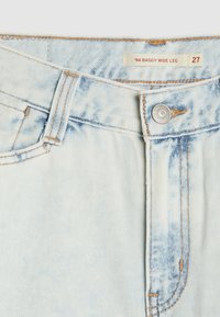 Šviesiai mėlyni džinsai su laisvu plačiu kojų dizainu, turintys sidabrinį užsegimą, kontrastingą siūlę ir etiketę su užrašu '94 BAGGY WIDE LEG 27'.