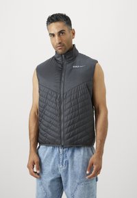 Gray sleeveless puffer vest with a high collar, zippered front, and horizontal quilted pattern on the torso; Nike logo and "BARCA" details.