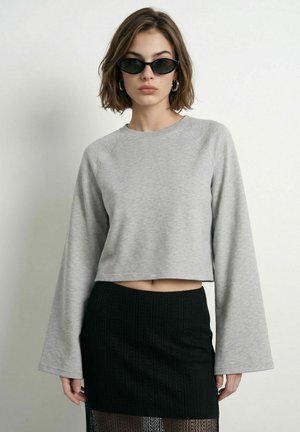 Femme portant des lunettes de soleil noires en forme d'œil de chat, un top court gris à manches longues, et une mini-jupe noire texturée, debout devant un fond uni.