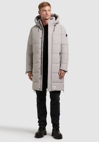 Cappotto imbottito grigio chiaro con cappuccio, caratterizzato da una chiusura zip frontale, tasche con zip e dettagli neri. Indossato sopra una maglietta nera e pantaloni scuri.