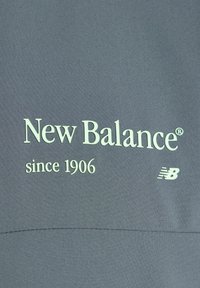 Logotipo de New Balance y texto "desde 1906" sobre un fondo de tela gris con un pequeño emblema "NB" a la derecha.