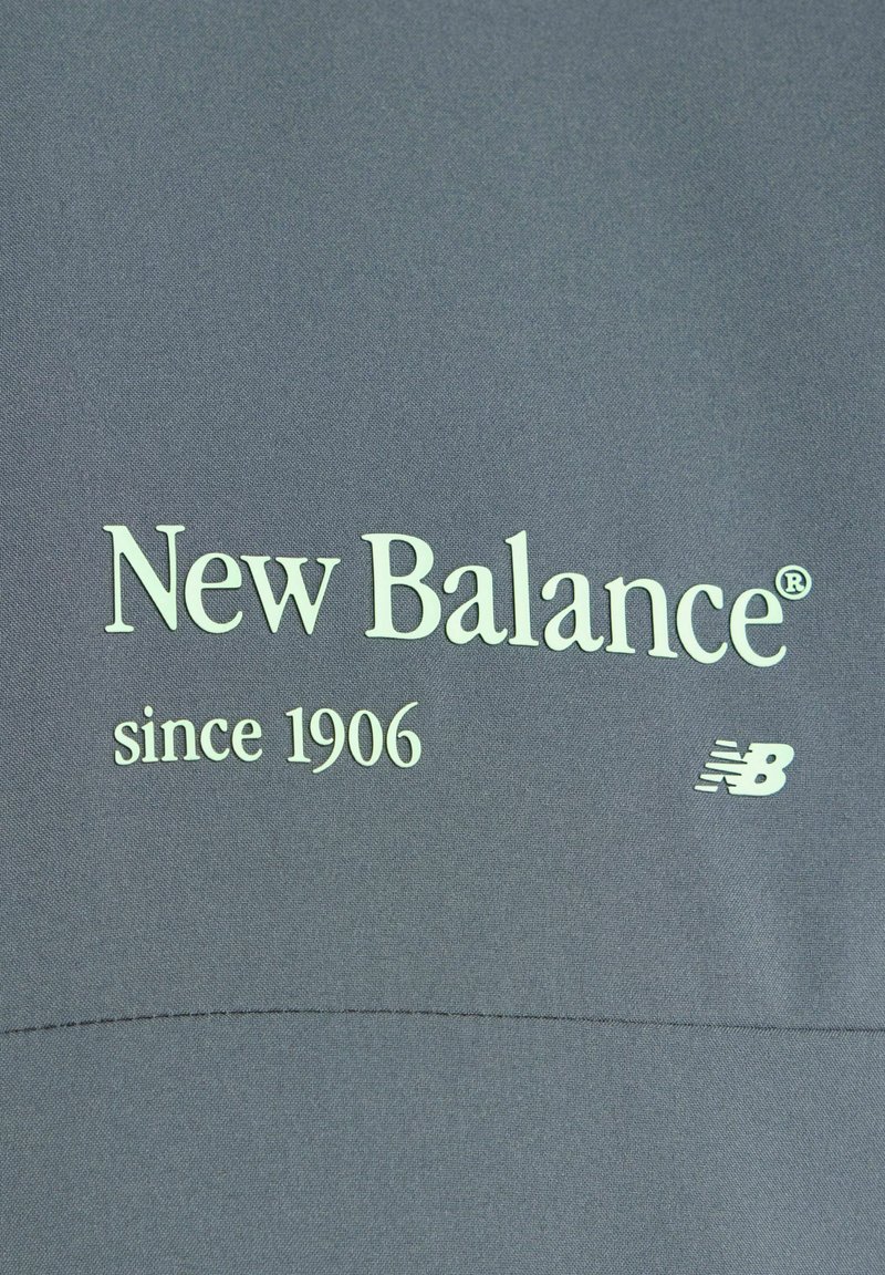 New Balance-logo en tekst "sinds 1906" op een grijze stofachtergrond met klein "NB" embleem aan de rechterkant.