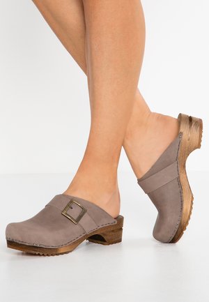 Sabots en suède gris avec un talon en bois, présentant une boucle en métal décorative et un design facile à enfiler, mettant en valeur une texture lisse et des lignes épurées.