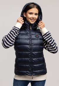 Granatowa kamizelka puchowa z wysokim kołnierzem i zamkiem błyskawicznym, wyposażona w naszywkę z logo, boczne kieszenie oraz pikowaną teksturę. Poniżej widoczny sweter z długim rękawem w paski.