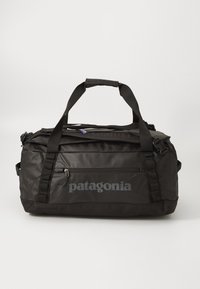 Crna Patagonia torba s ručkama na vrhu, prednjim zip džepom i podesivim remenom za rame na jednostavnoj pozadini.