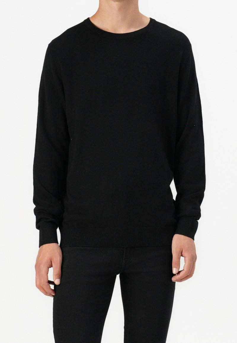 Homme portant un pull noir uni à manches longues et un pantalon noir, debout devant un fond blanc.