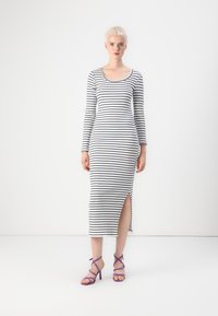 Pasiasta sukienka z długim rękawem w białym i czarnym prążkowanym materiale z bocznym rozcięciem. Modelka nosi purpurowe sandały na szpilce. Minimalistyczne tło.