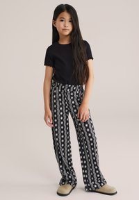 Crna rebrasta majica kratkih rukava u kombinaciji s hlačama širokih nogavica s uzorkom koje sadrže bijele valovite crte i dizajne sunčevog izbočenja, upotpunjeno bež cipelama bez petice.