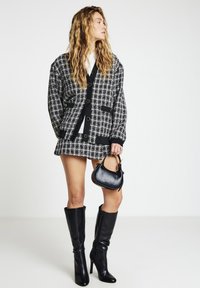 Veste en tweed noir et blanc avec des épaules structurées, associée à une mini-jupe assortie, des bottes noires à talons hauts et un petit sac à main noir.