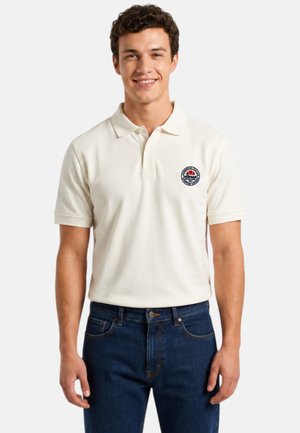 Jeune homme aux cheveux courts, bouclés et foncés, portant un polo blanc avec un petit logo rond et un jean bleu, souriant et debout.