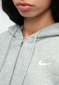 Tuvojums personai, kura valkā gaiši pelēku Nike džemperi ar rāvējslēdzēju, redzami lūpas, zods, kakls un melni mati.