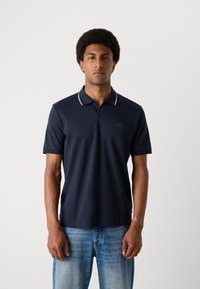 Ναυτικά μπλε πόλο μπλουζάκι από μαλακό ύφασμα με αντίθετο γιακά και μικρό λογότυπο στο στήθος. Κοντά μανίκια και άνετη γραμμή.