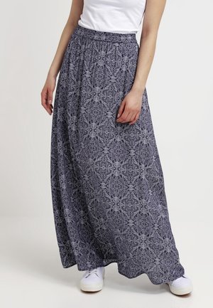 Kobieta ubrana w długą, granatową spódnicę w wzory z elastycznym paskiem, białe trampki oraz białą, bezrękawnikową bluzkę włożoną do spódnicy.