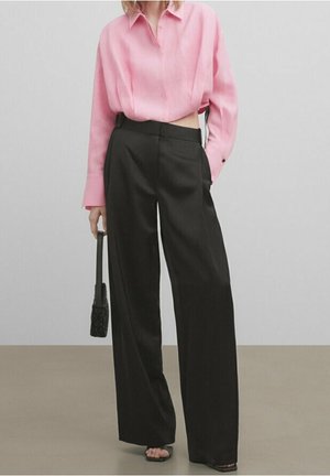 Person wearing a loose pink long-sleeve shirt tucked into high-waisted wide-leg black pants, holding a small black handbag.