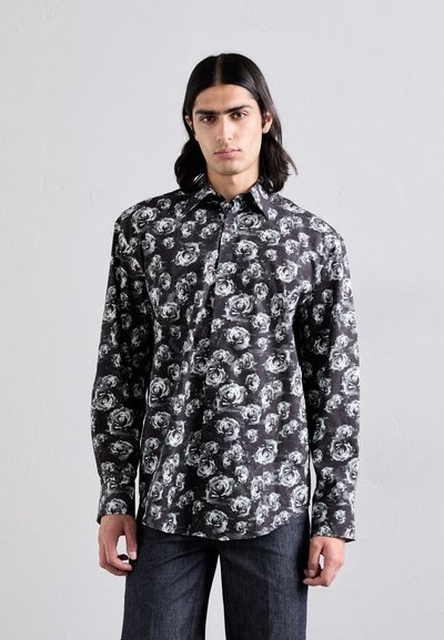 Chemise à manches longues en tissu noir, ornée d'un motif floral blanc. Design à boutonner avec un col et un ourlet droit. Portée avec un jean noir.
