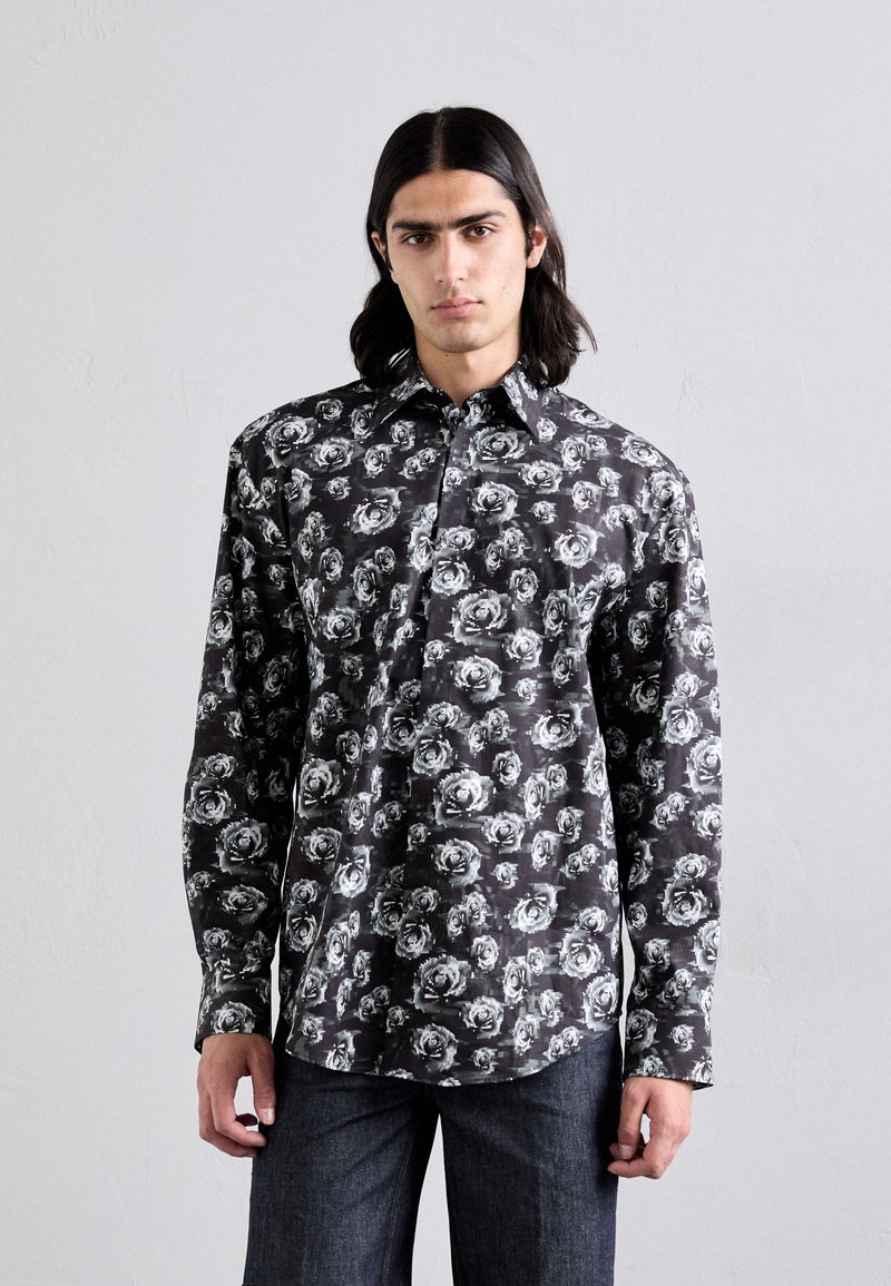Long-sleeve shirt in black fabric, featuring a white floral pattern. Button-up design with a collar and straight hem. Worn with dark jeans.