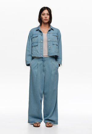 Kobieta stojąca, ubrana w jasnoniebieską kurtkę dżinsową i szerokie spodnie z beżową bluzką oraz brązowe sandały na białym tle.