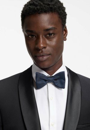 Ung mand iført en sort smokingjakke, hvid skjorte og blå mønstret butterfly mod en ensfarvet hvid baggrund.