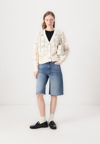 Kremfarget strikket cardigan med åpenmønster, svart topp, lyseblå denim Bermuda-shorts, hvite sokker og svarte loafers. Liten kremfarget håndveske.