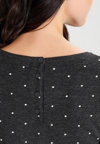 Szary sweter z dzianiny w białe kropki, z okrągłym dekoltem z tyłu i trzema czarnymi guzikami przy szyi. Miękka tekstura, casualowy design.