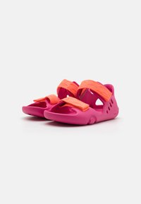 Pink children's sandals featuring thick orange Velcro straps, open design, textured rubber material, and ventilation holes along the sides.