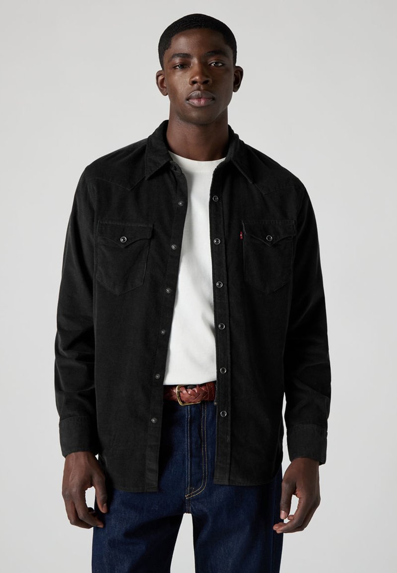 Camisa preta de veludo cotelê com gola pontiaguda, fecho de botões à frente, dois bolsos no peito e tecido texturizado, usada sobre uma t-shirt branca e jeans de ganga.