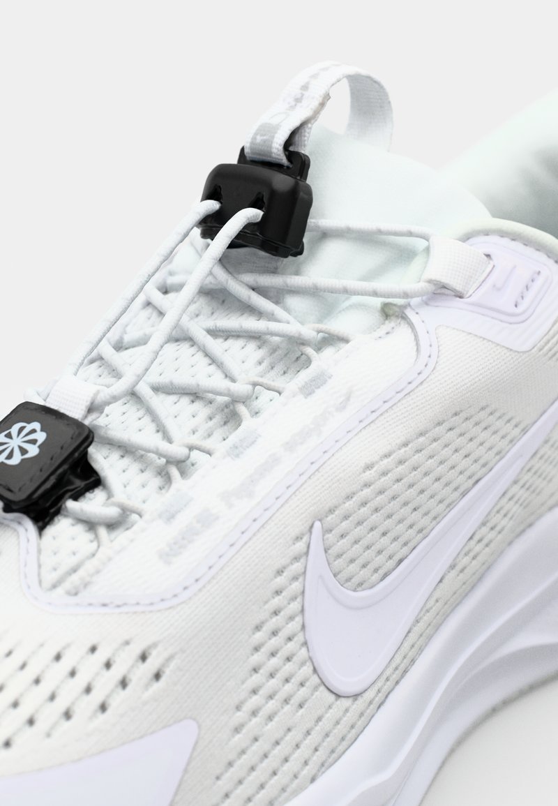 Zapatilla deportiva blanca con parte superior de malla, paneles texturizados, cordones elásticos y un mecanismo de bloqueo negro. Presenta un logo de swoosh blanco.