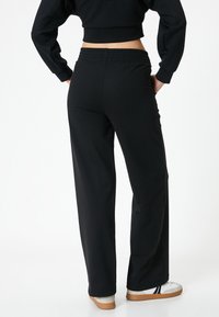 Schwarze, elastische weite Hose mit glatter Textur und weichem Fall. Kombiniert mit weißen und beigefarbenen Schuhen.