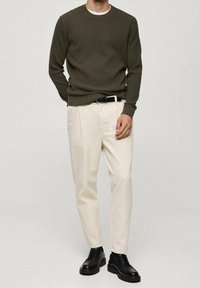 Uomo che indossa un maglione a coste verde scuro, pantaloni beige con pieghe e cintura nera, e scarpe stringate in pelle nera, posando su uno sfondo neutro.