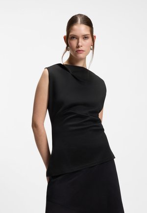 Femme aux cheveux bruns attachés, portant des boucles d'oreilles en or en forme de anneaux et un haut noir sans manches, debout devant un fond blanc uni.