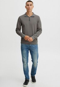Sivi zip-up dukserica s ovratnikom, dugim rukavima i rebrastim manžetama, u kombinaciji s plavimfarmerkama i crnim tenisicama na neutralnoj pozadini.