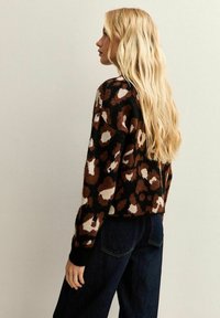 Vrouw met lang blond haar, gekleed in een bruine, zwarte en witte luipaardprint trui en donkerblauwe jeans, zijwaarts kijkend tegen een effen muur.