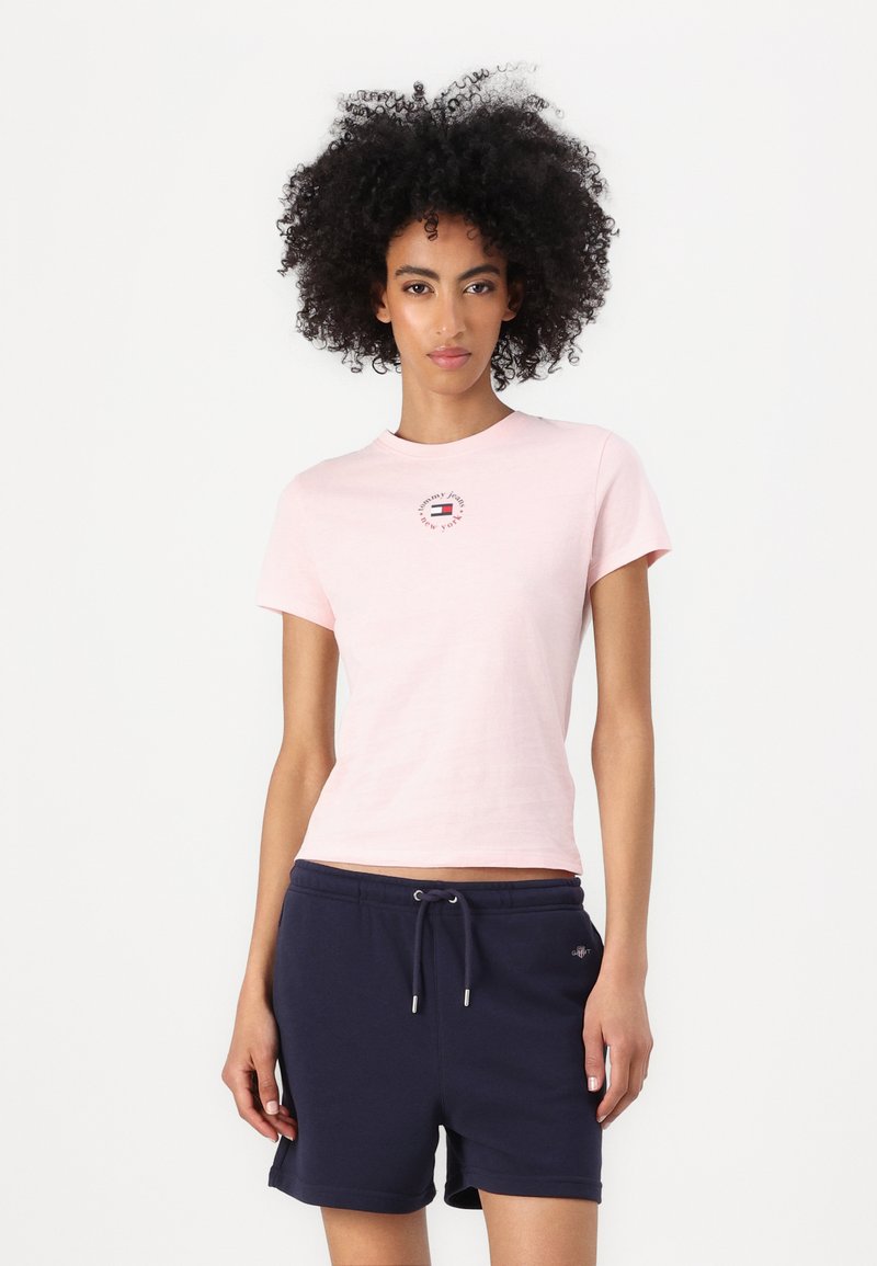 Light pink cotton t-shirt with short sleeves and a round neckline, featuring a small printed logo on the front. Paired with dark blue shorts.
