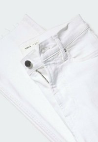 Jean en denim blanc avec fermeture éclair visible, bouton, passants de ceinture et ourlets effilochés, plié sur une surface blanche.