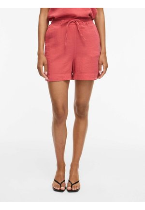 Kvinde iført koralrosa teksturerede shorts med bundet taljebælte og sorte åbne sandaler står mod en hvid baggrund.