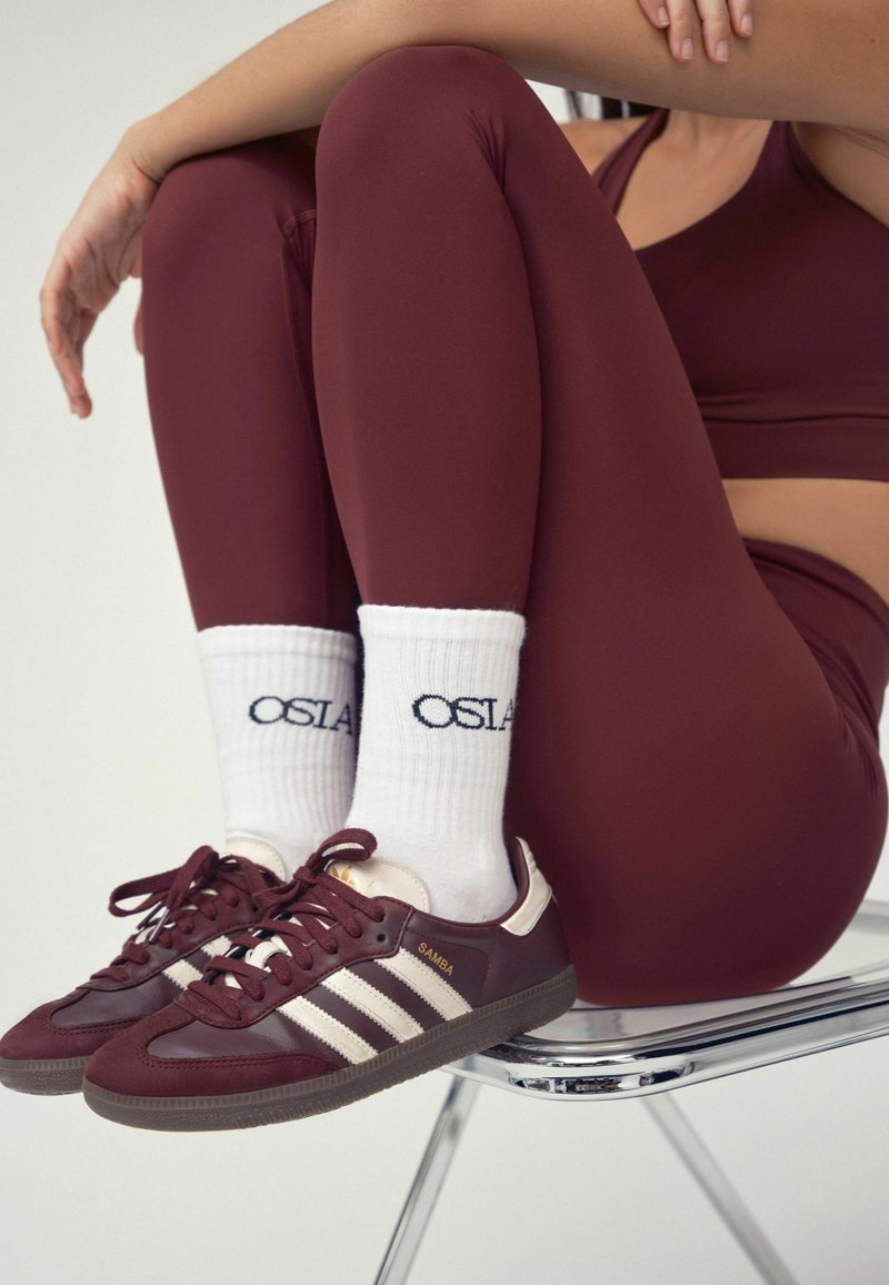 Personne portant des leggings bordeaux et un soutien-gorge de sport, des chaussettes blanches avec texte et des baskets Adidas Samba bordeaux, assise sur une chaise à cadre métallique.