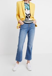 Geel blazer, gestreepte zwart-witte top, lichtblauwe wijde jeans en witte sneakers. De blazer heeft twee knopen en een ontspannen pasvorm.