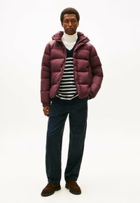 Giacca puffa bordeaux con cappuccio, polsini elastici e finitura lucida, indossata sopra un maglione a righe e pantaloni in velluto a coste scuri. Mocassini marroni.