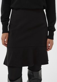Black flared skirt made of soft material, features a smooth texture with a waistband and a ruffled hem. Paired with black tights and boots.