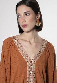 Abito marrone con tessuto texturizzato, scollatura a V, bordatura decorativa ricamata e chiusura con bottoni. Gli accessori includono grandi orecchini in perline.
