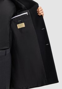 Cappotto di lana con esterno nero e fodera nera liscia. Presenta una tasca con zip e un'etichetta che identifica il materiale come lana pura.