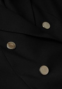 Tissu noir avec trois boutons ronds argentés brillants disposés en diagonale sur le vêtement, montrant le détail des coutures et de la texture.
