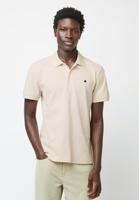 Polo beige in cotone, con un piccolo logo a forma di albero blu navy sul petto, maniche corte e un classico design del colletto.