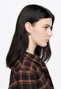 Camisa de cuadros negra y naranja con cuello; el modelo lleva pendientes pequeños en forma de aro. El cabello es liso y oscuro, cayendo por debajo de los hombros.